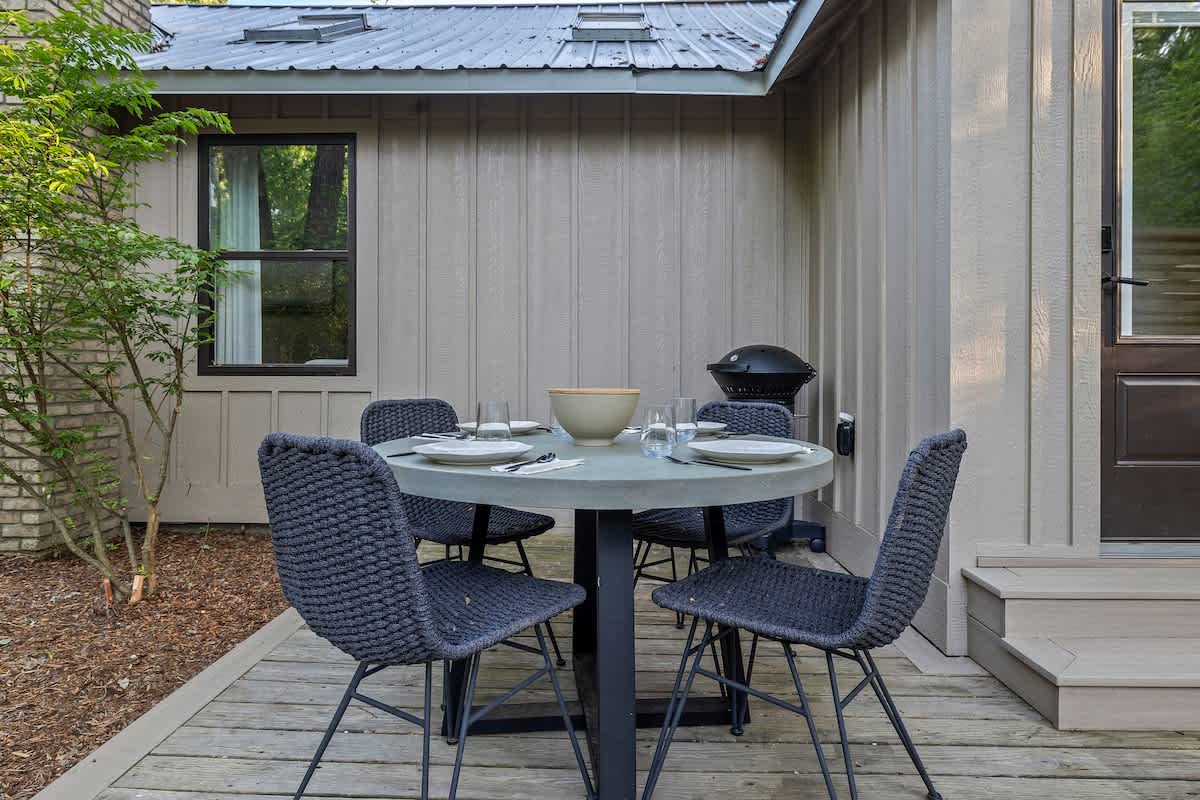 A private outdoor deck features a modern round dining table with four dark woven chairs, set with plates and glasses, next to the light-colored siding of a cottage with a window, and surrounded by wood chips and greenery.