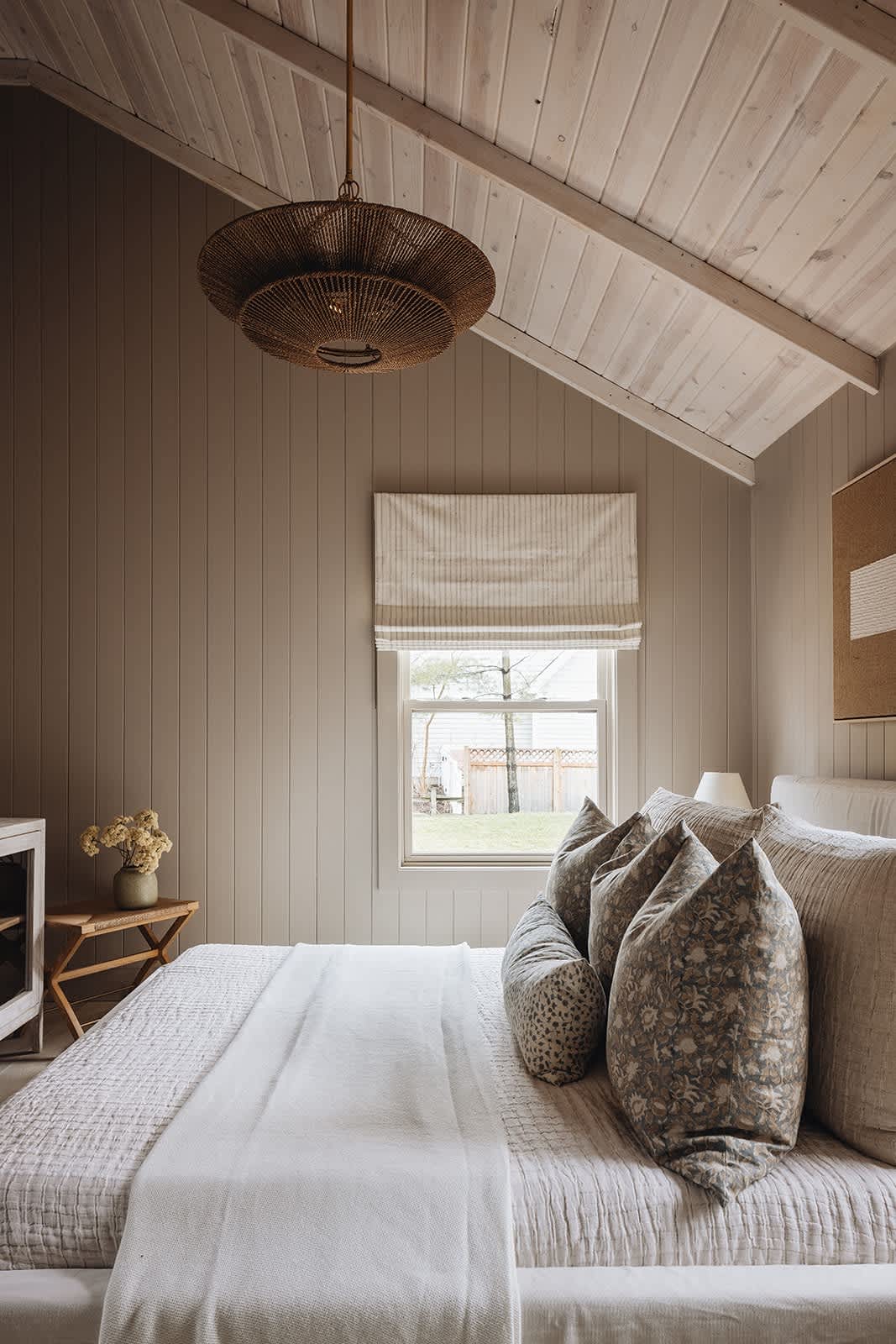View of the bedroom with a large bed, a window featuring a striped Roman shade, and a small wooden side table with a vase of dried flowers, all under a vaulted wood-paneled ceiling.