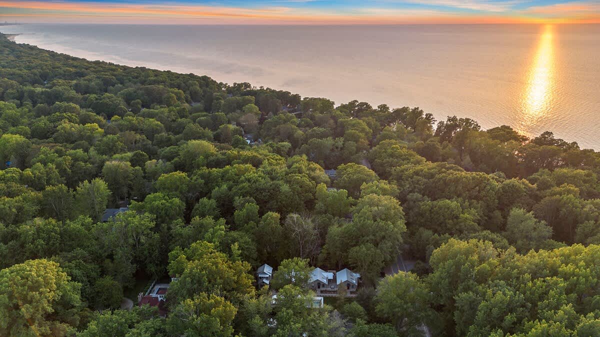 An aerial view showcases a dense green forest landscape with scattered houses, leading to a vast body of water under a colorful sunset sky. A bright sunbeam reflects on the water's surface, highlighting the serene coastal setting.
