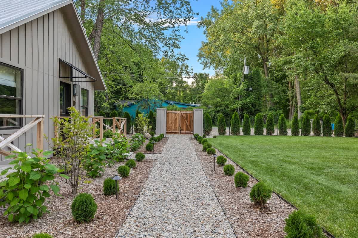 A long gravel path, bordered by small green shrubs and landscape lighting, extends past a modern farmhouse-style cottage on the left, leading towards a wooden gate and a line of tall evergreen trees, with a vibrant green lawn on the right.