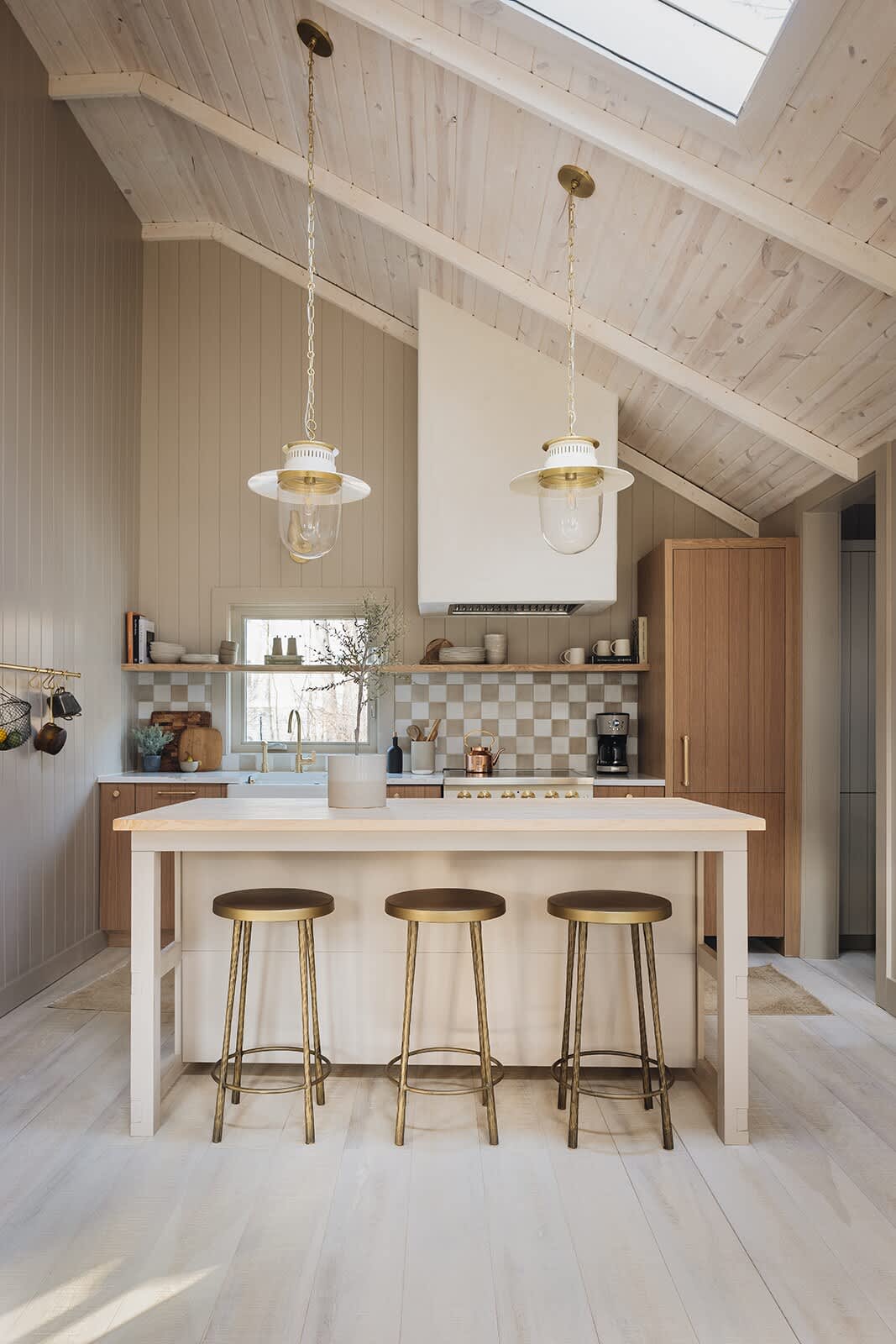 A bright kitchen with a vaulted wood-paneled ceiling and skylight features a large light wood island with three brass bar stools. Two glass and brass pendant lights hang above, illuminating the light wood cabinetry and checkered tile backsplash.