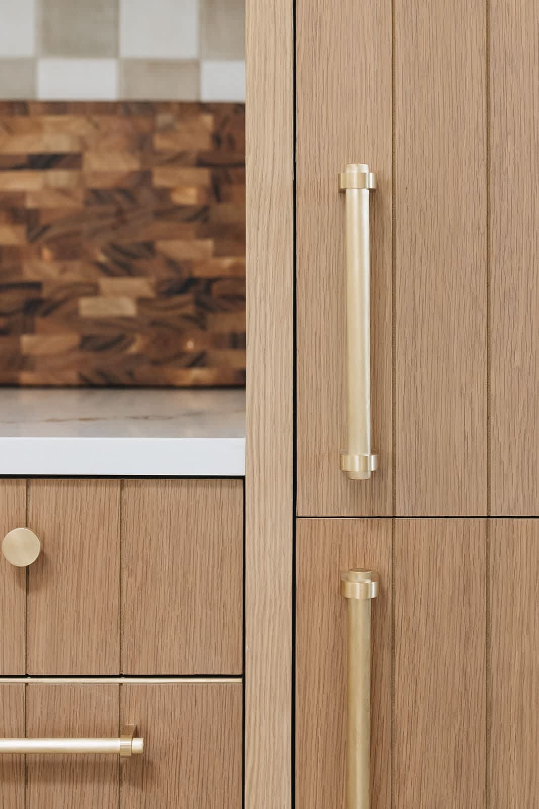 Detail shot of light wood kitchen cabinetry with vertical paneling, featuring sleek brass pull handles and a round brass knob, with a wooden cutting board partially visible in the background.