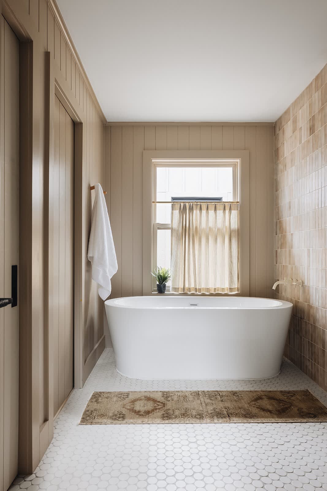 Bright bathroom featuring a modern freestanding white bathtub, a window with a sheer curtain, and warm-toned rectangular wall tiles, complemented by a patterned bath mat.