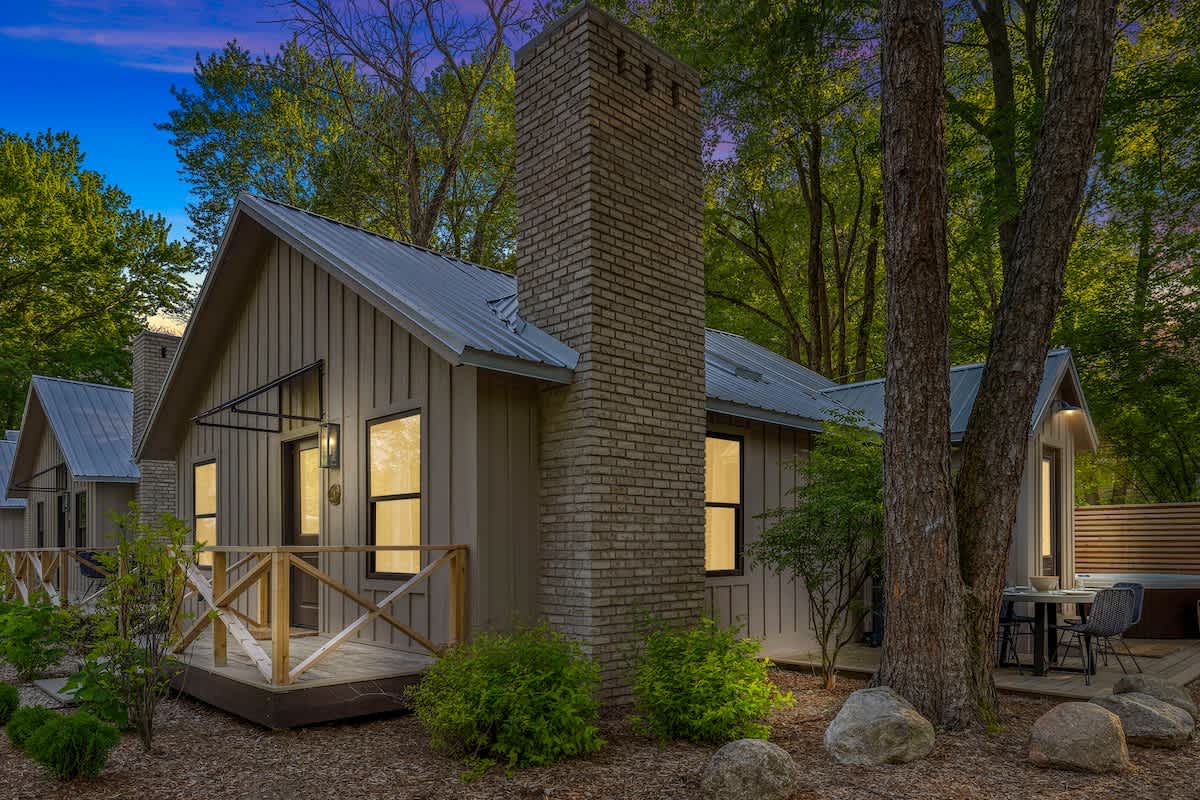 A modern farmhouse-style cottage with light siding, a metal roof, and a prominent brick chimney is nestled among tall trees, featuring a small wooden deck with railings and illuminated windows, suggesting a cozy evening ambiance.