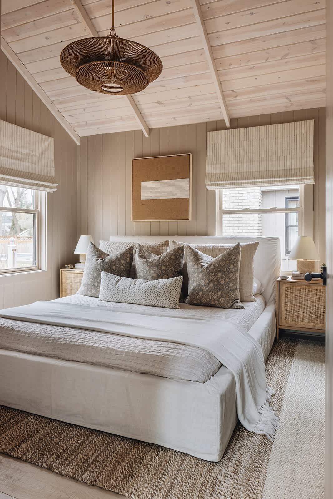 Serene bedroom with a large bed, two cane nightstands, and windows with striped Roman shades, all beneath a vaulted white-washed wood ceiling with a woven pendant light fixture.