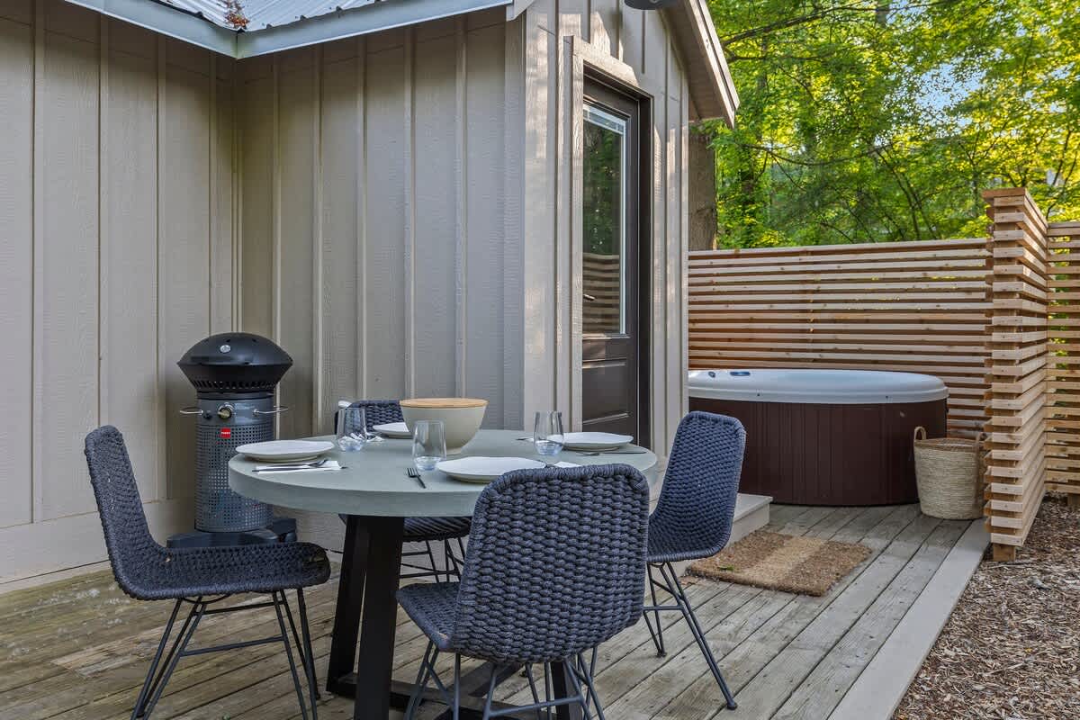 A private outdoor deck features a round hot tub enclosed by a slatted wooden privacy screen, a modern round dining table with four woven chairs, and a black BBQ grill, all set against the light siding of the cottage.