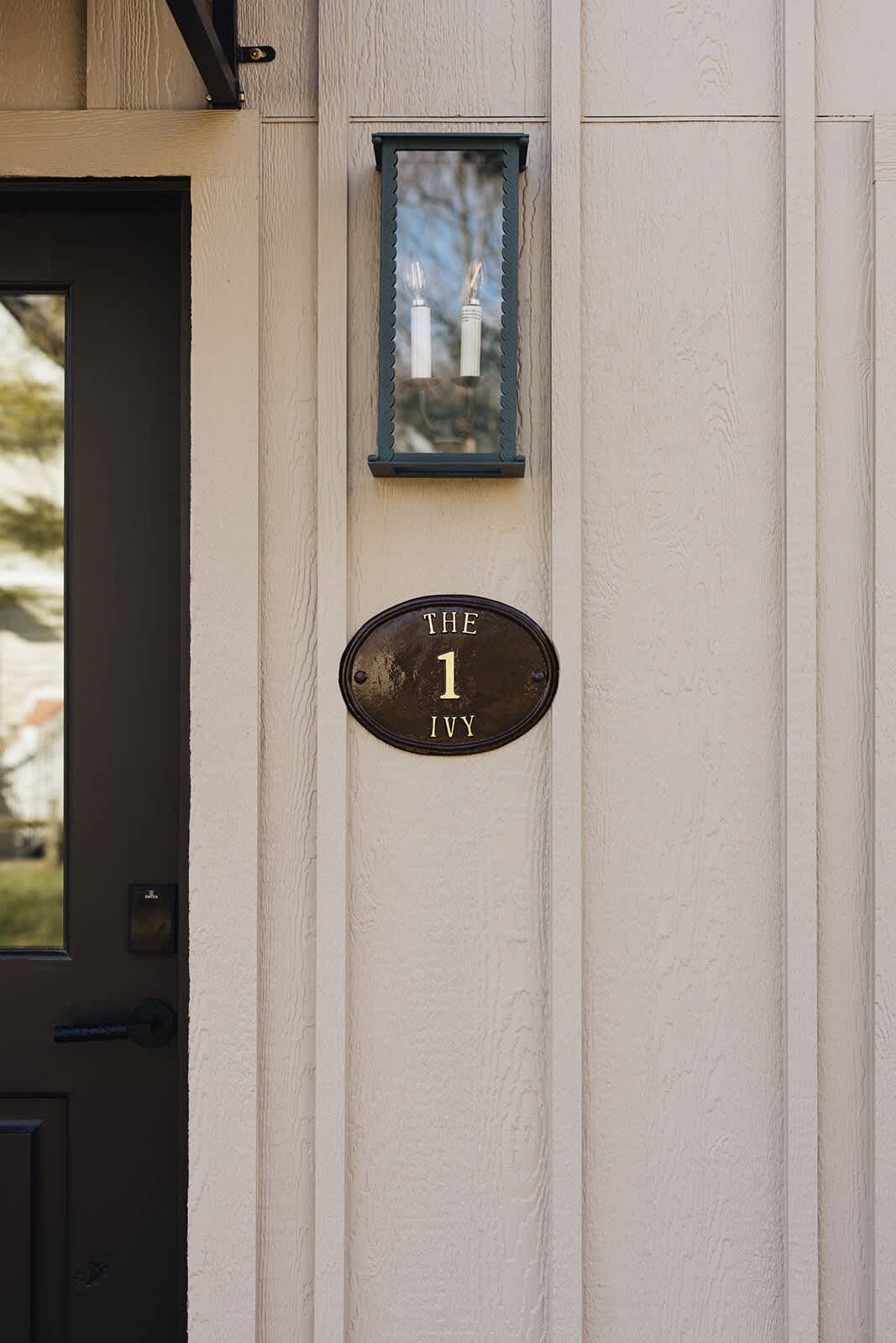 Close-up of the front entrance of a cottage, showing light beige vertical siding and a dark front door with a small black awning. A decorative oval plaque reads "THE IVY 1", illuminated by an outdoor lantern.