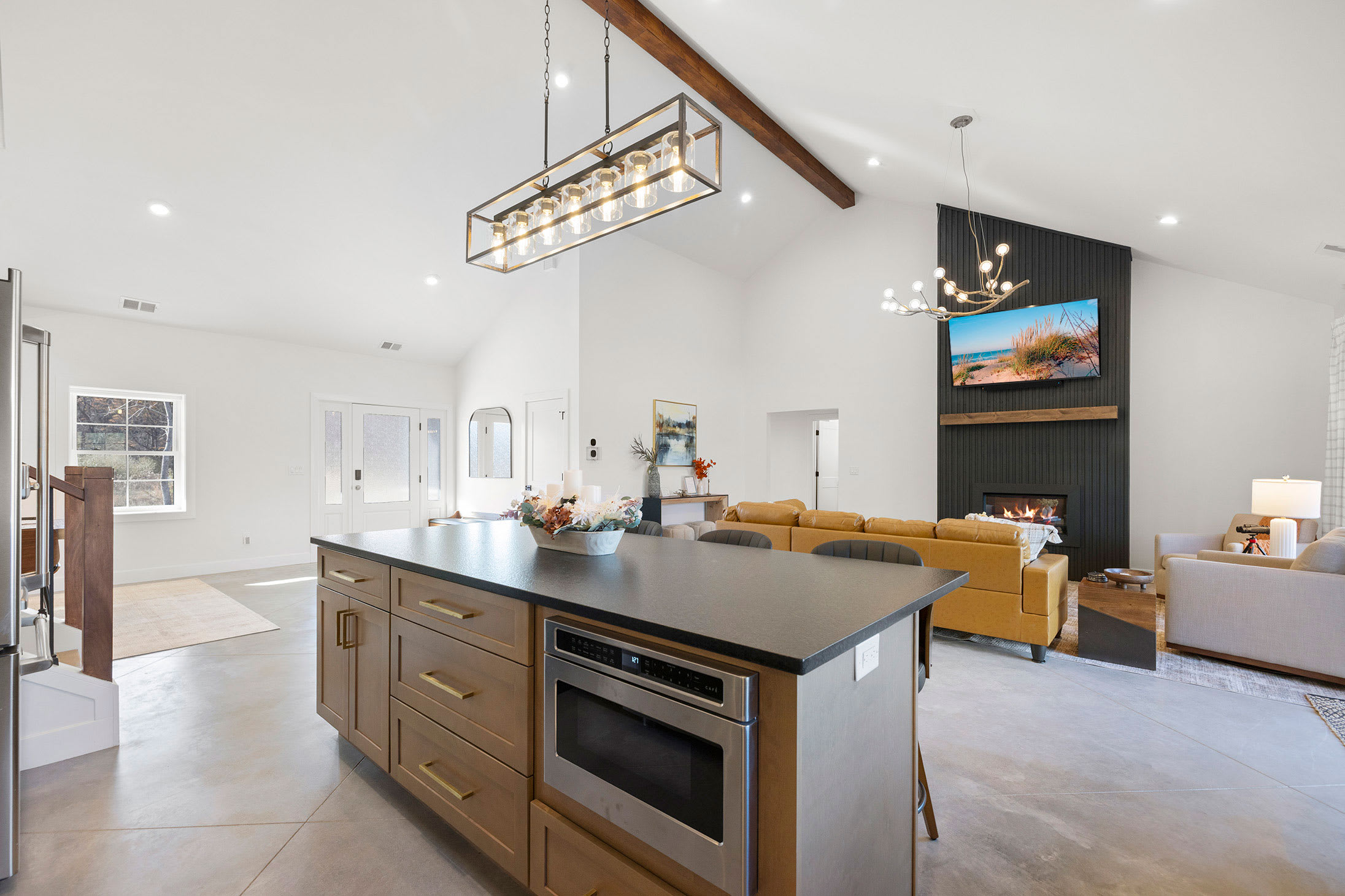 Expansive open-concept living area with a vaulted white ceiling, exposed wooden beam, a large kitchen island, and a modern fireplace with a black shiplap accent wall and a mounted TV.