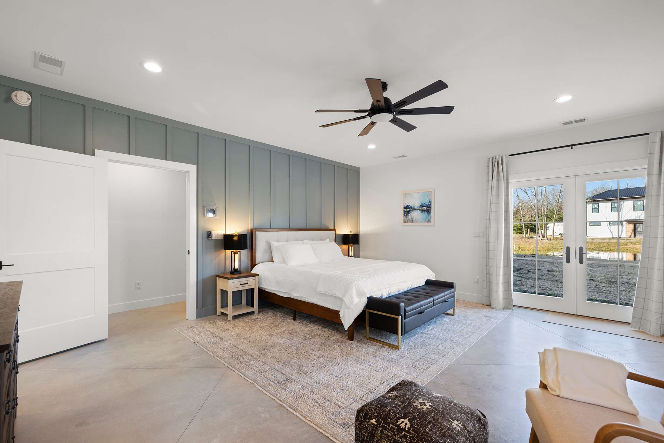Spacious bedroom with a light grey accent wall behind a white upholstered bed, dark wood nightstands with modern lamps, a ceiling fan, and French doors leading to the outside.