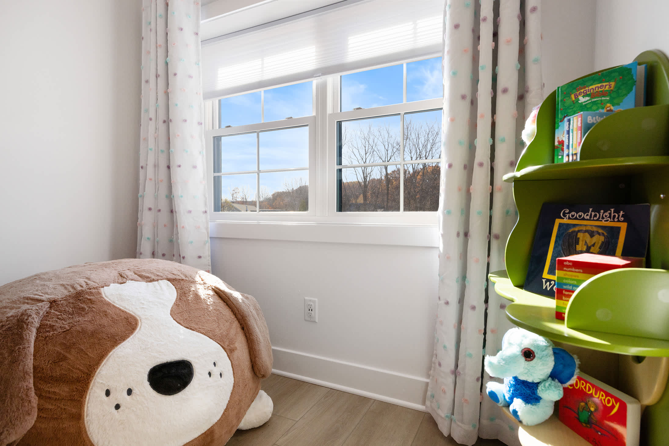 A bright child's room featuring a large window with white blinds, colorful polka-dot curtains, a plush dog-shaped beanbag, and a green bookshelf filled with children's books and toys.