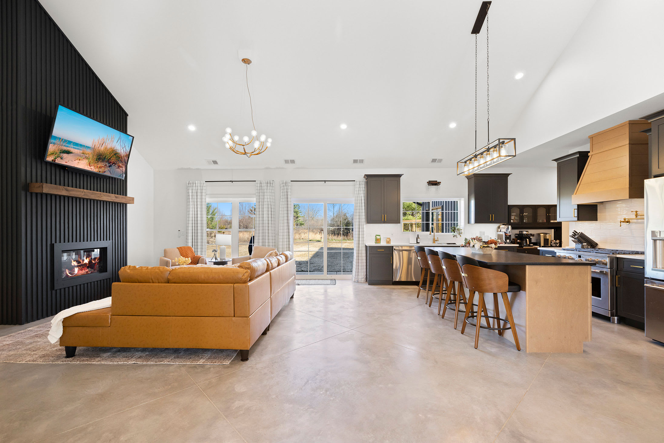 Wide view of the open-concept living space, featuring a tan leather sectional facing a modern black shiplap fireplace with a mounted TV, and an adjacent kitchen with dark cabinetry and a large island with bar stools.