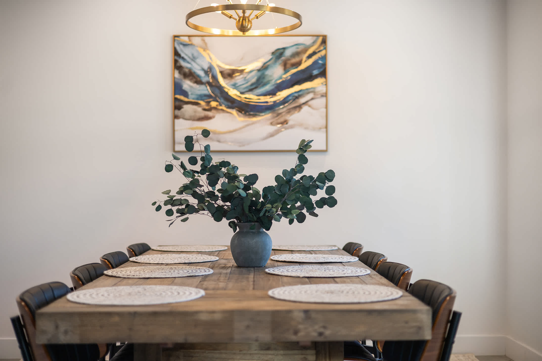 Close-up of the rustic wooden dining table set with placemats and a centerpiece of eucalyptus branches in a grey vase, beneath a modern gold chandelier and abstract wall art.