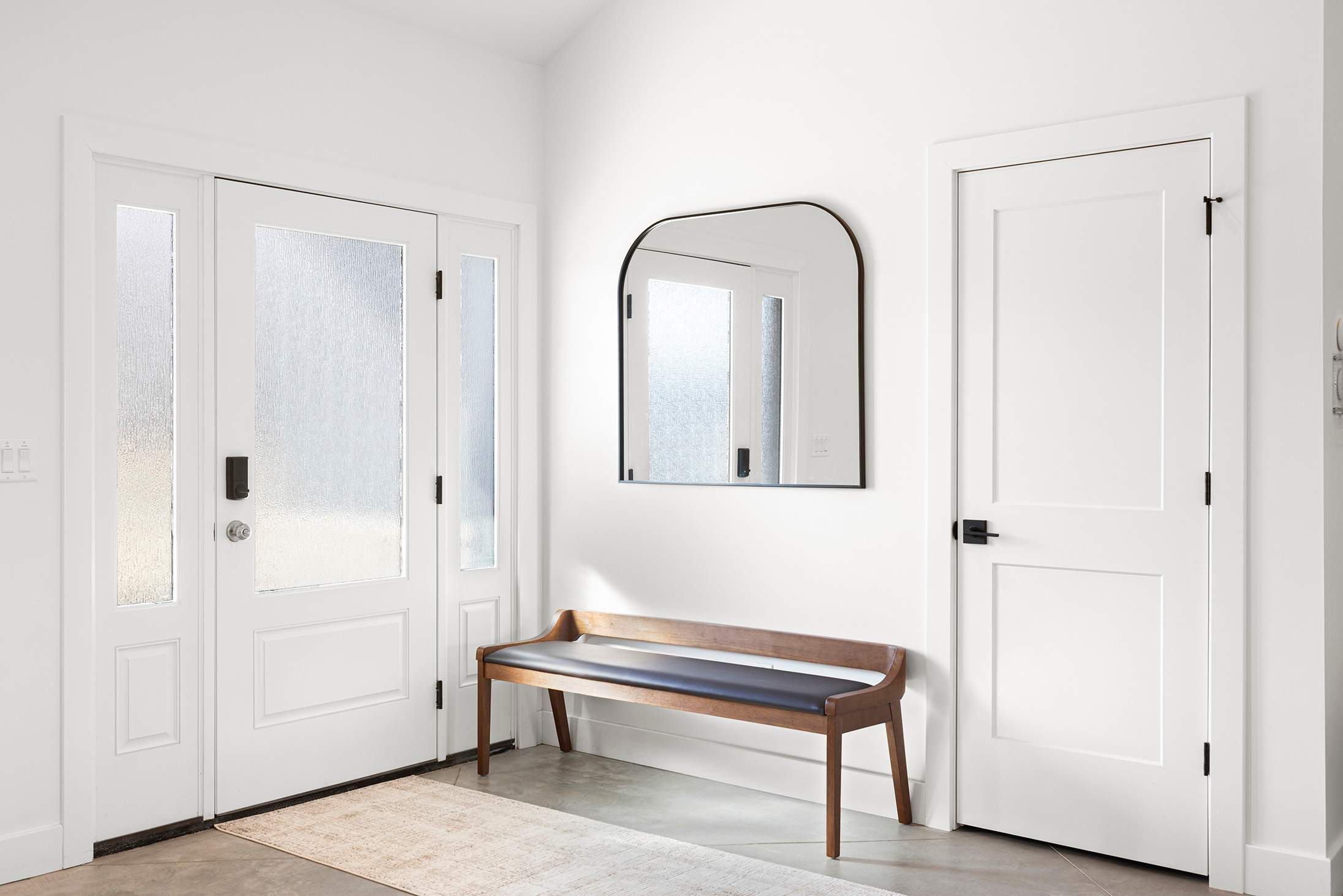 Bright entryway with a modern black-framed arched mirror above a wooden bench with a black cushion, flanked by a white front door with frosted glass and a solid white interior door.