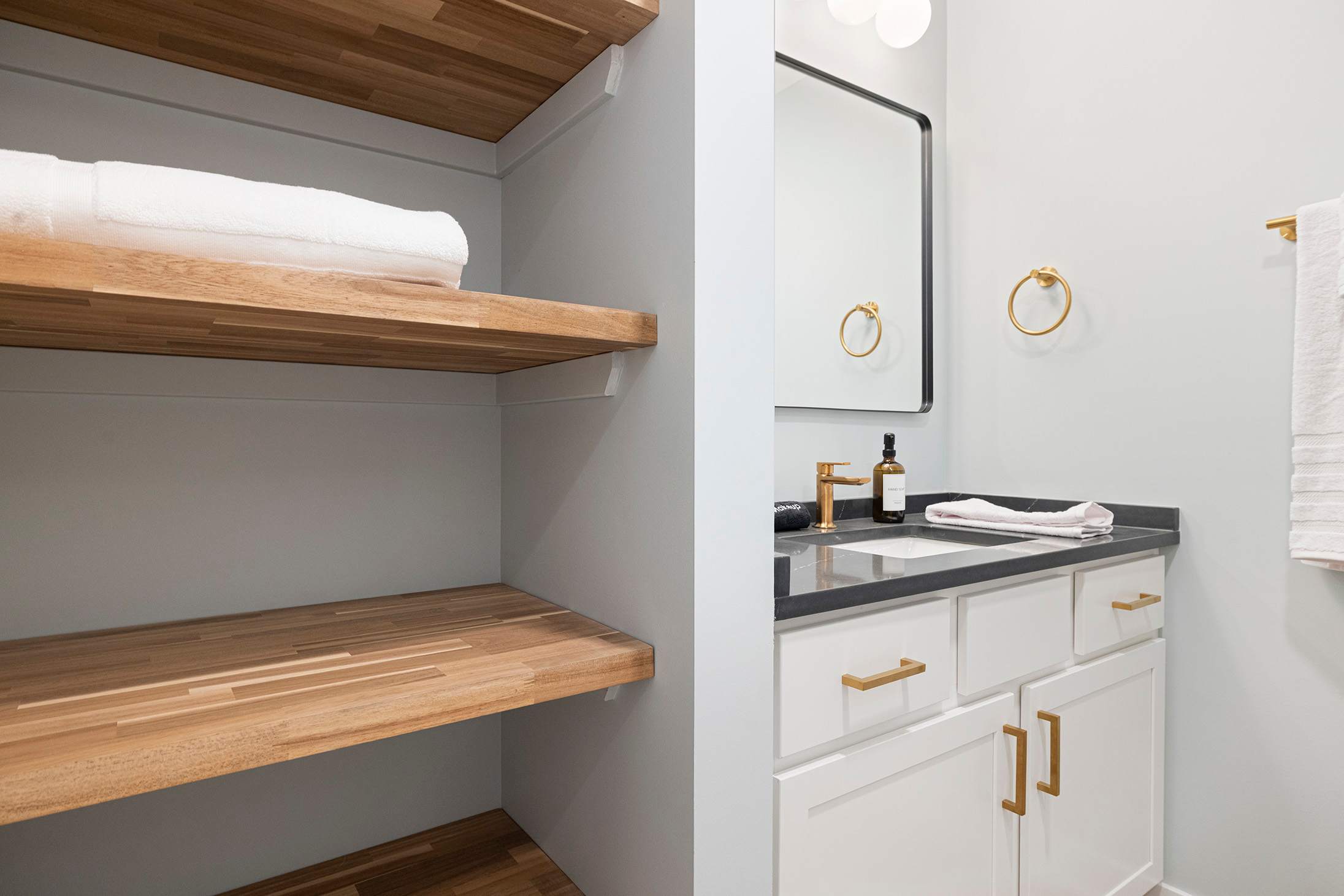 A detailed view of a modern bathroom vanity with white cabinets, a dark countertop, gold faucet and hardware, a rectangular mirror, and open wooden shelving with a rolled white towel.