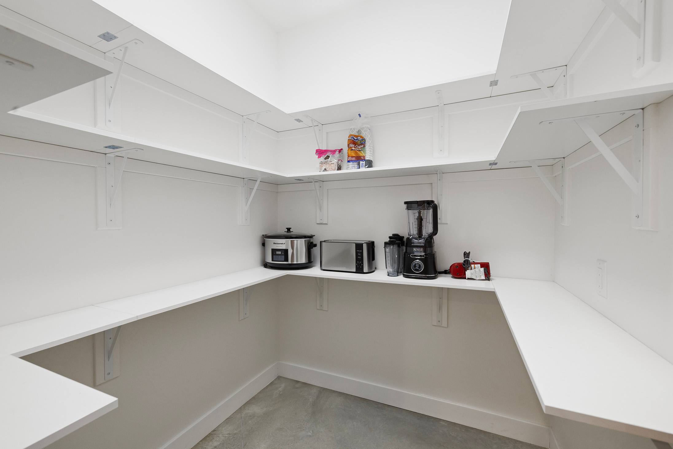 A well-organized walk-in pantry with white built-in shelves stocked with food items and small kitchen appliances like a slow cooker, toaster oven, and blender.