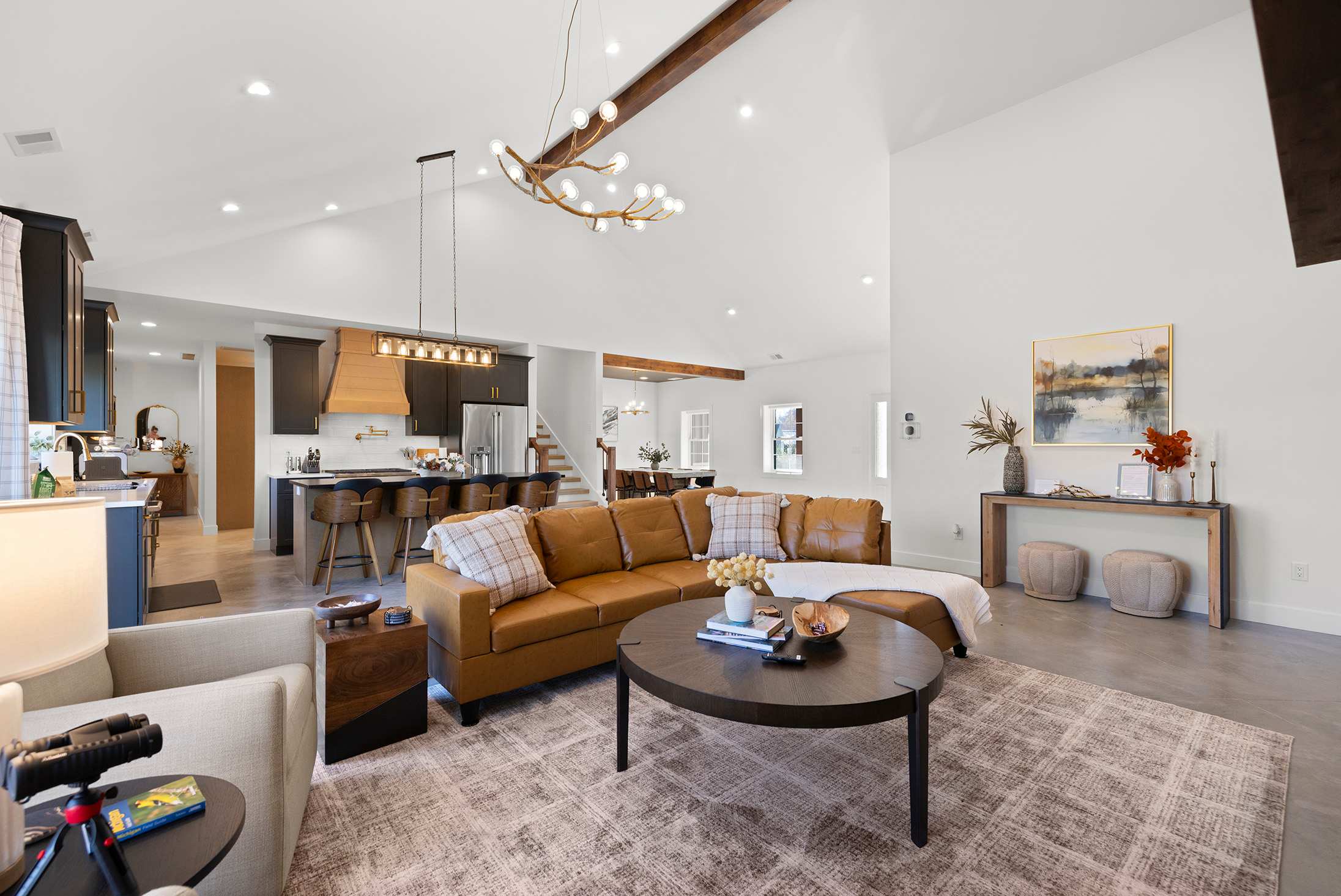 Expansive open-concept living area with a large brown leather sectional sofa, a round coffee table, and a view into the modern kitchen with dark cabinetry and a large island, all under a vaulted ceiling.