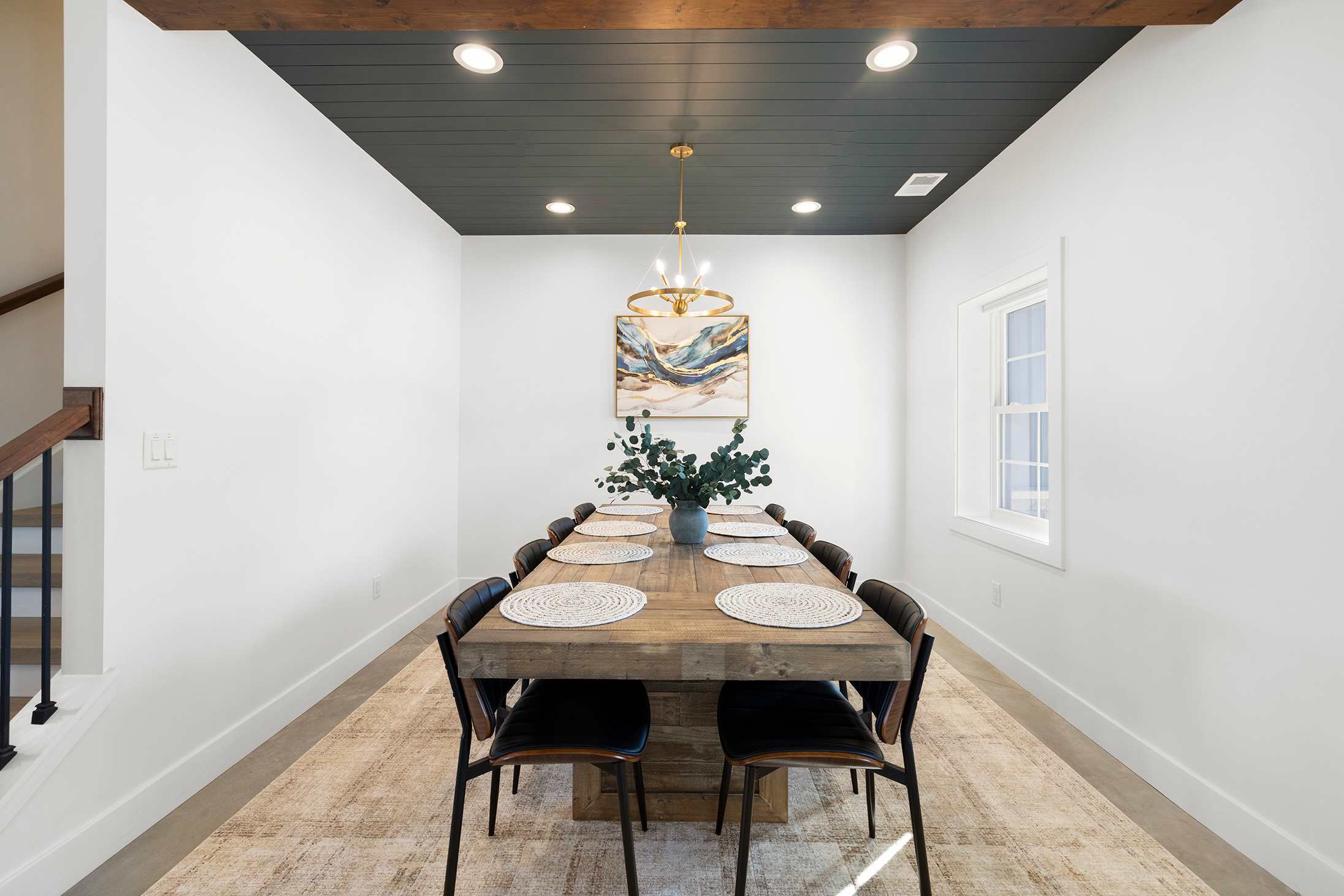 Elegant dining room with a long wooden table, black leather chairs, a dark shiplap ceiling, and a modern gold chandelier, complemented by abstract art on the white wall.