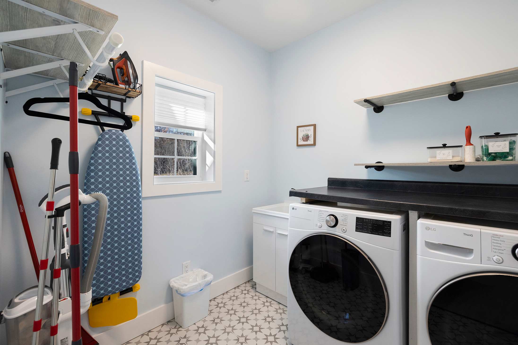 Functional laundry room with light blue walls, a white washer and dryer, a utility sink, patterned floor tiles, and shelving for cleaning supplies including an ironing board and iron.