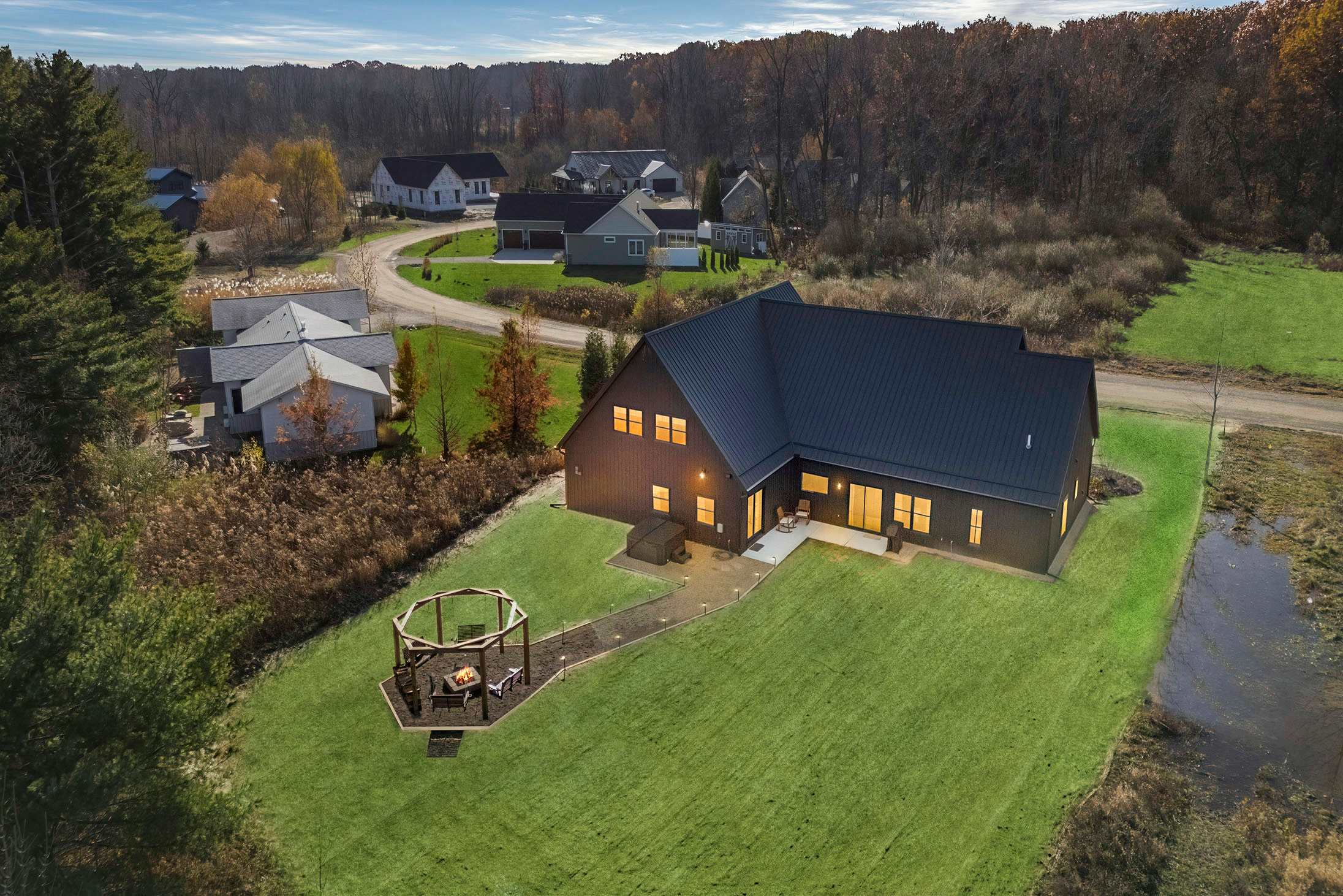Aerial view showcasing the back of the modern home with a dark metal roof and siding, a concrete patio, a fire pit pavilion in the grassy yard, and surrounding natural landscape.