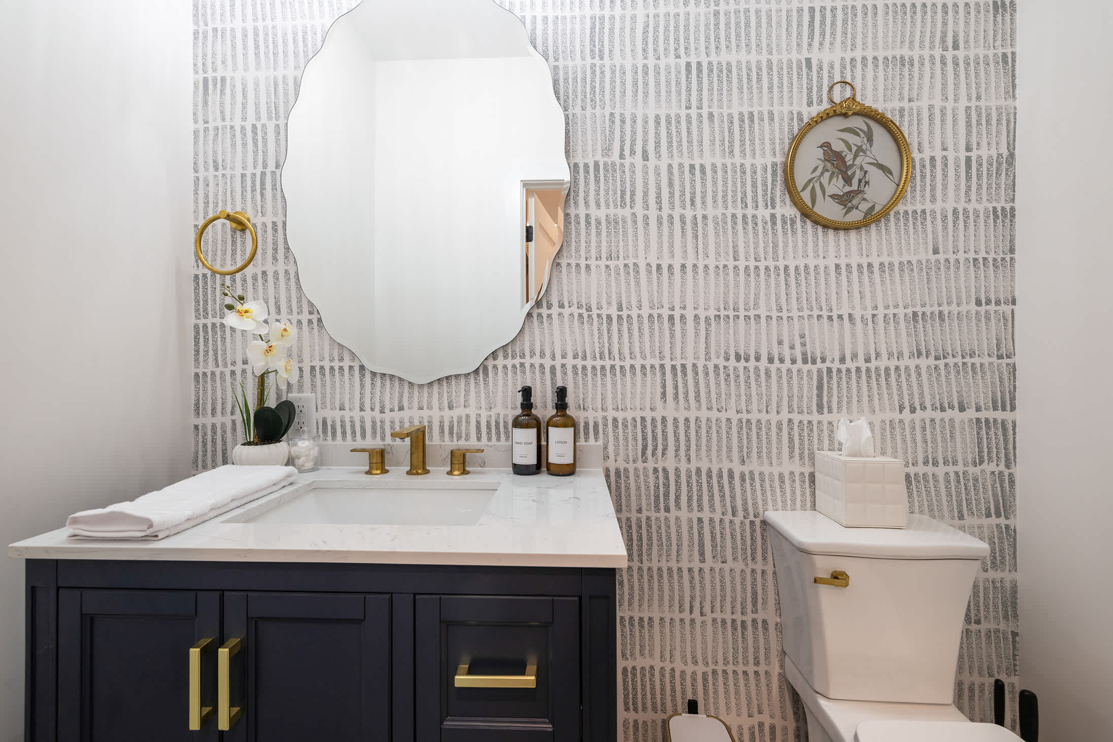 Stylish powder room featuring a white marble-top vanity with a dark blue cabinet and gold fixtures, a unique scalloped mirror, and a striking grey and white patterned wallpaper.
