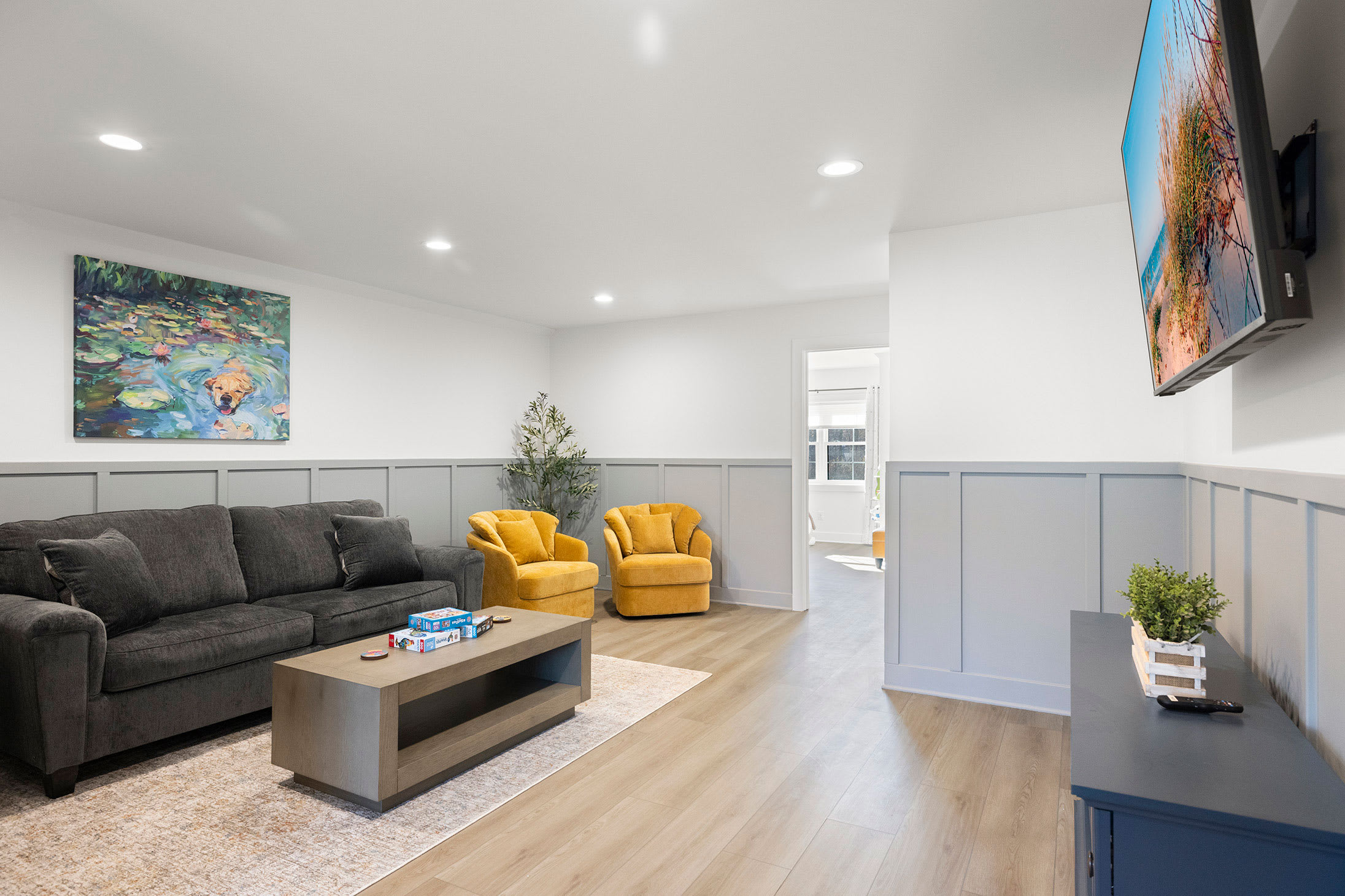 Cozy living area with a dark grey sofa, two vibrant yellow armchairs, a light wood coffee table with board games, and a wall-mounted TV, all set against white walls with light grey wainscoting.