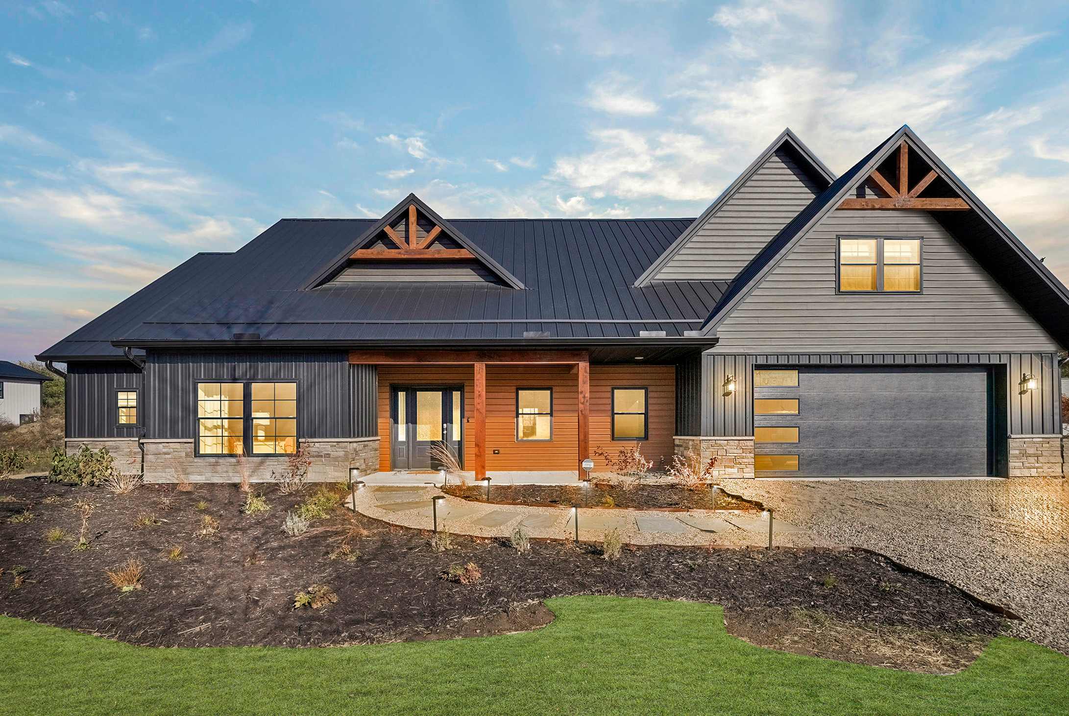 The front exterior of a modern farmhouse-style home with dark siding, a dark metal roof, natural wood accents around the entrance, and a garage door with horizontal windows.