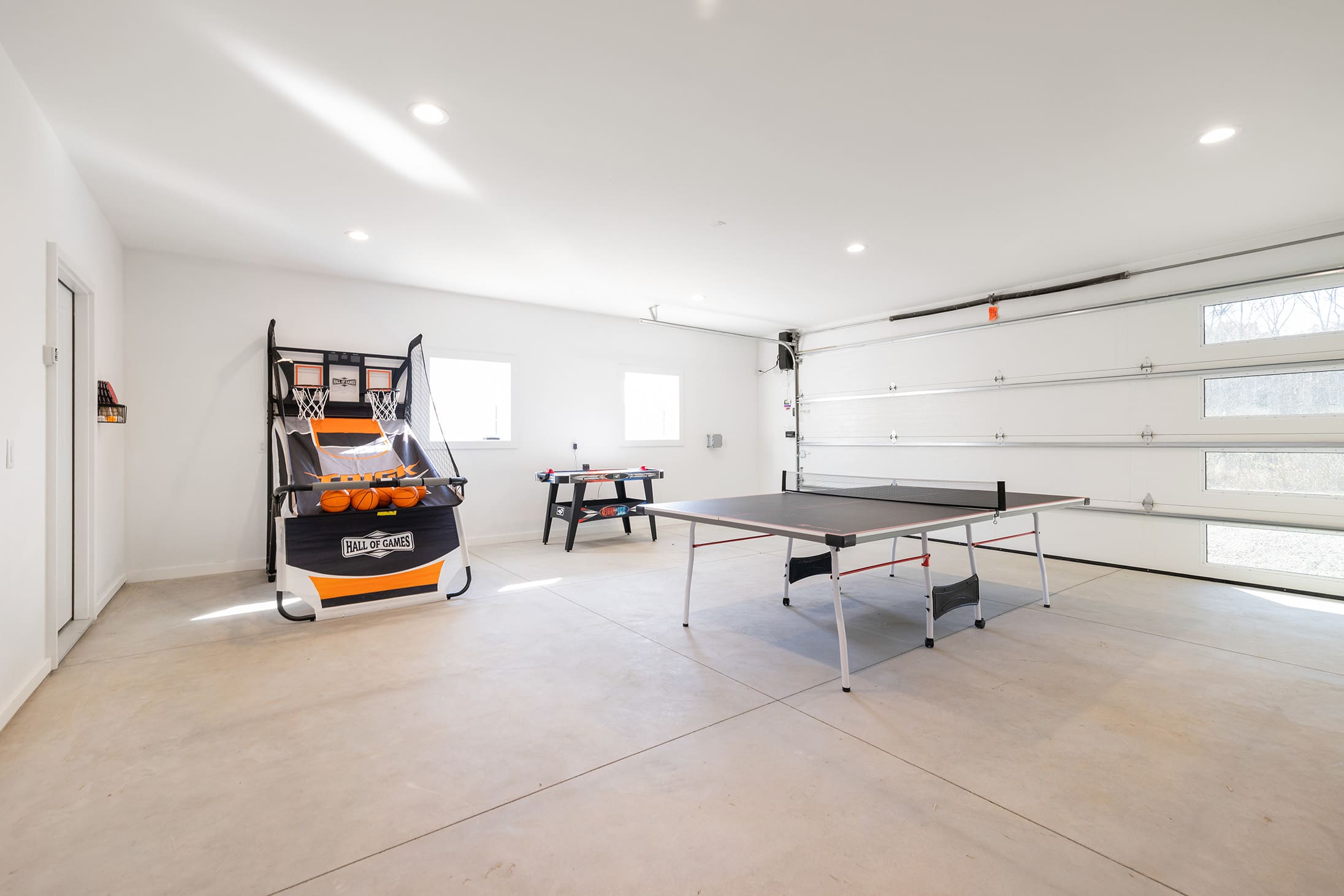 Large game room set in a garage space with a concrete floor, featuring a ping pong table, an arcade basketball game, and a smaller foosball table, with a white garage door visible.
