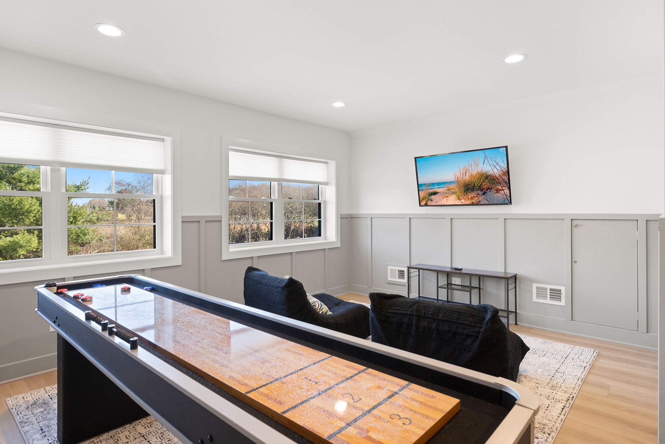 Bright game room featuring a large wooden shuffleboard table, two black bean bag chairs, a wall-mounted TV, and multiple windows, with light grey wainscoting along the walls.