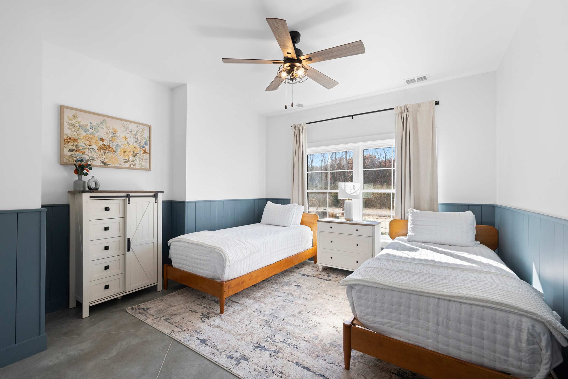 Bright and airy bedroom featuring two wooden twin beds with crisp white bedding, a white dresser with a barn door style, and a ceiling fan, set against white walls with a blue wainscoting.