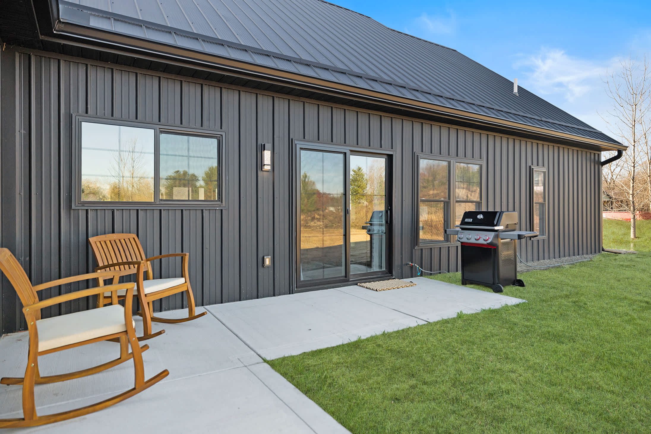 The back exterior of the house features dark vertical siding, a concrete patio with two wooden rocking chairs, and a BBQ grill, overlooking a grassy yard.