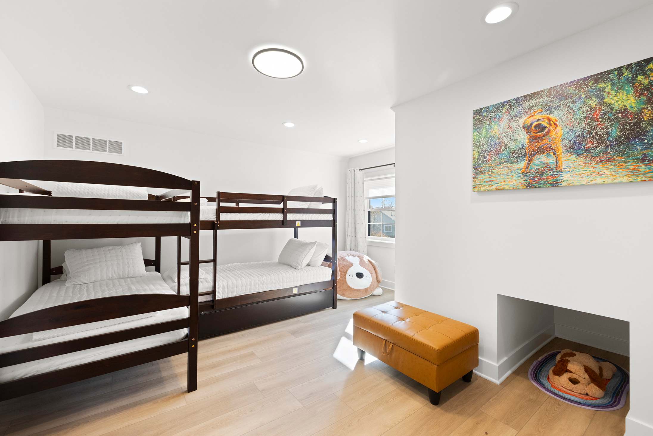Playful kids' bedroom with two dark wood bunk beds, a colorful dog painting above a decorative fireplace nook, and a brown leather ottoman, all on light wood flooring with a window.
