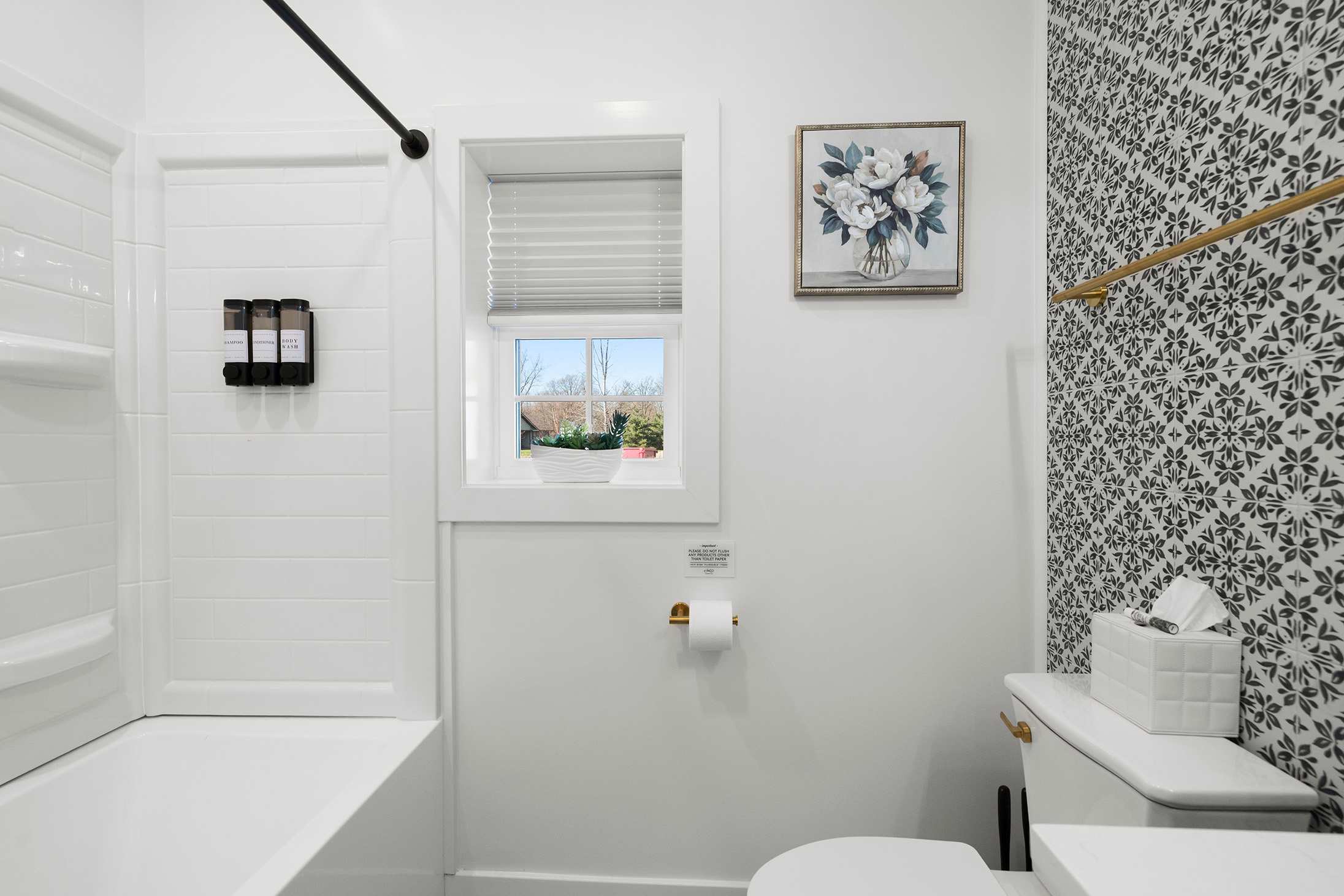 Clean bathroom with a white toilet, a shower/tub combo with white subway tiles, and a small window, complemented by a black and white patterned accent wall and floral artwork.