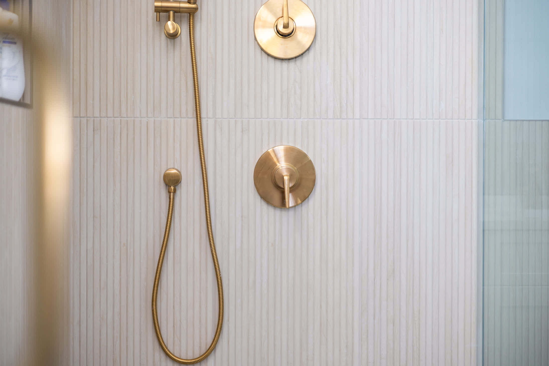 Close-up of modern gold shower fixtures, including a handheld showerhead and control knobs, set against a textured light-colored tiled wall inside a glass-enclosed shower.