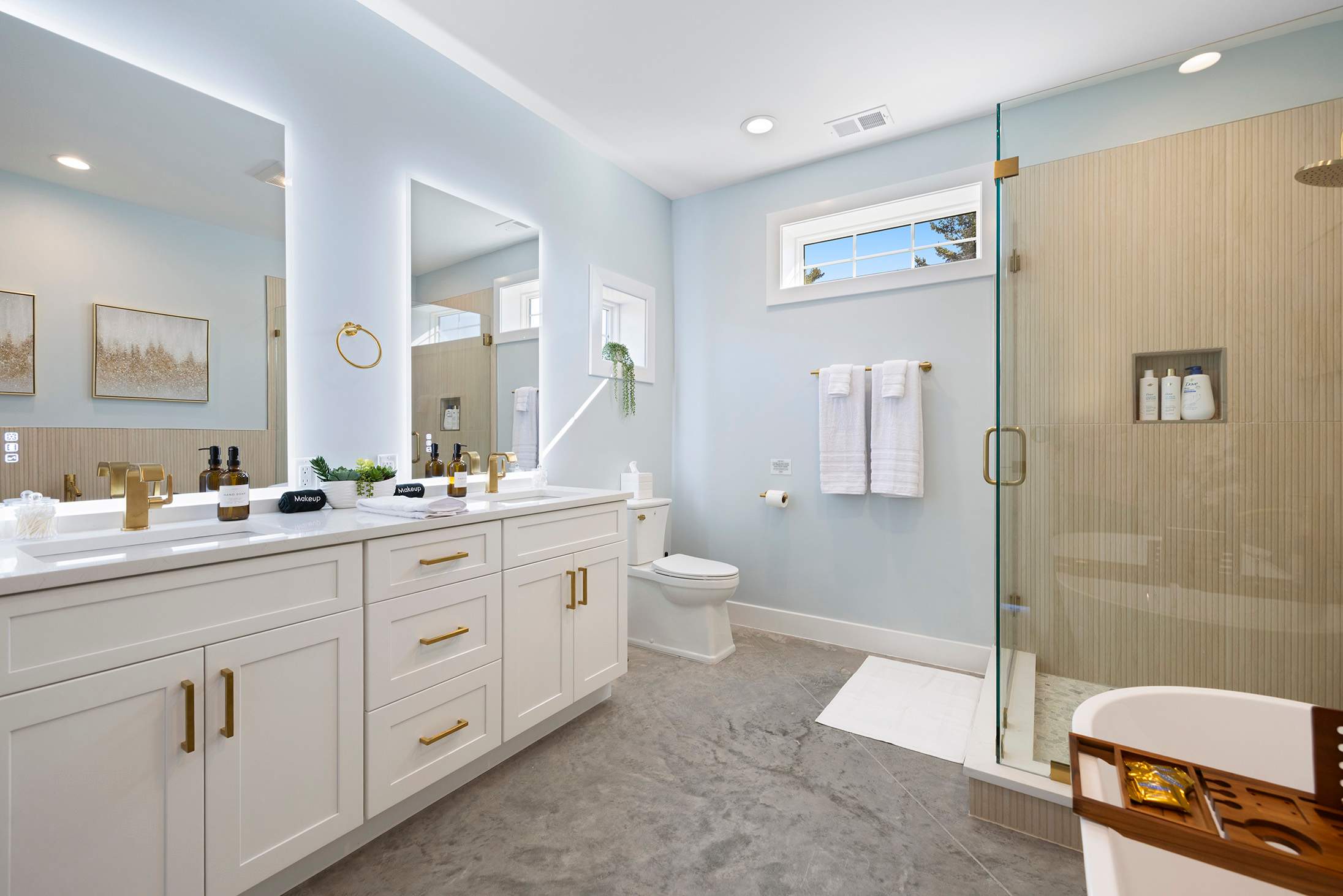 Bright modern bathroom featuring a double vanity with white cabinets, gold hardware, illuminated mirrors, a glass-enclosed shower, and a freestanding bathtub, all with light blue walls and grey floor tiles.