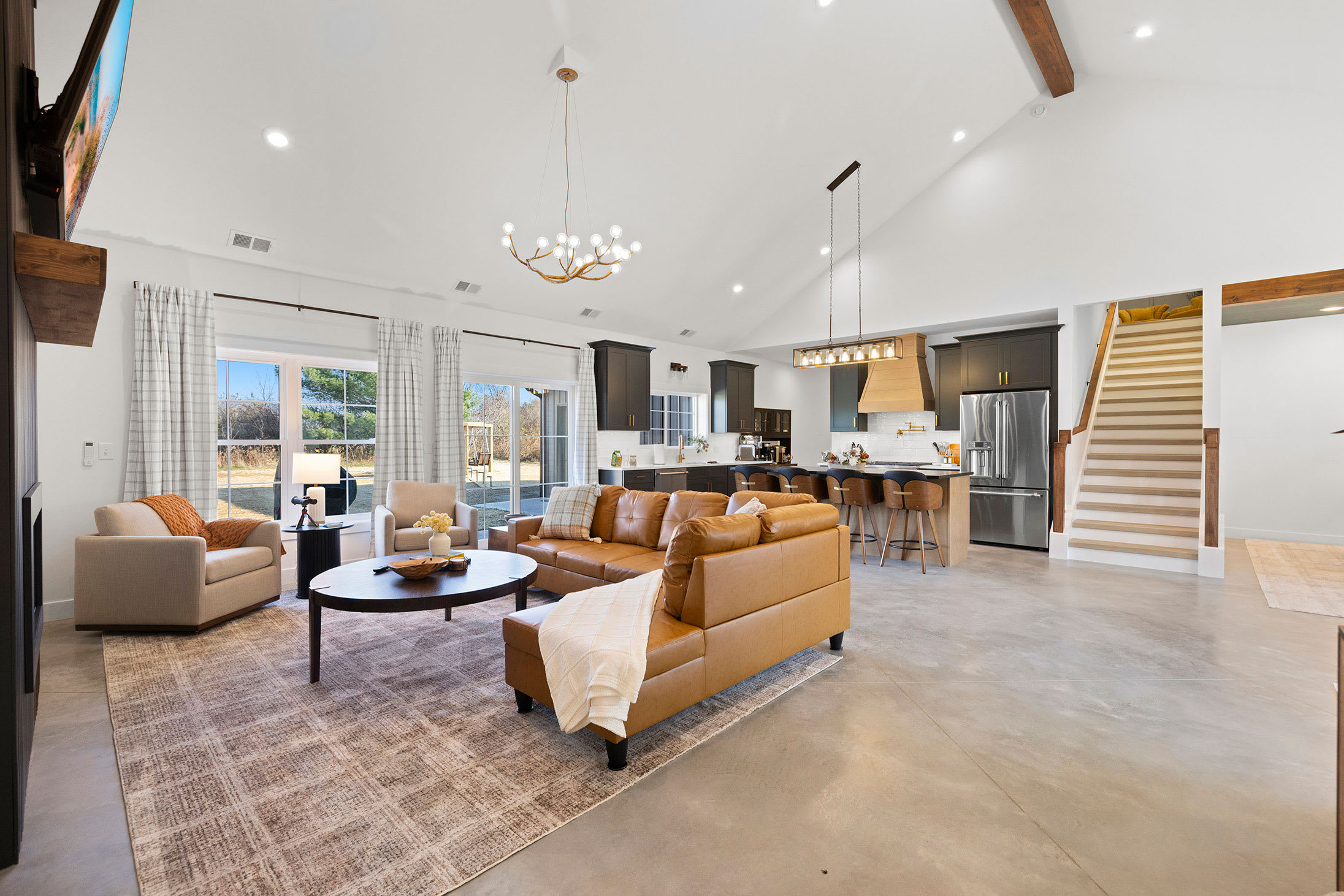 Bright open-concept living room with a large tan leather sectional sofa, a patterned area rug, and a view into the modern kitchen with dark cabinets and a large island. Sliding glass doors lead to the exterior.