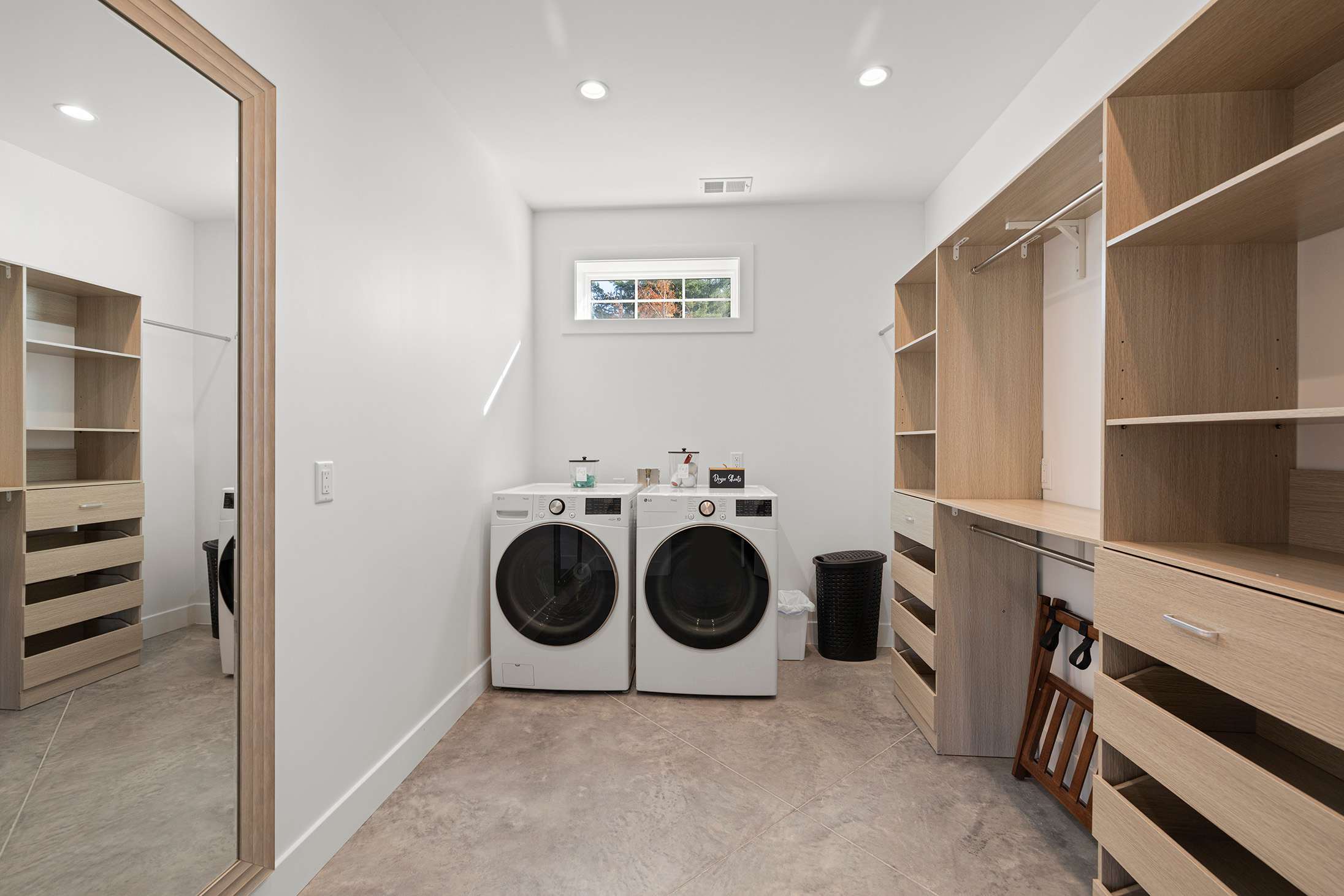 Spacious laundry room combined with a walk-in closet, featuring a washer and dryer, light-colored built-in shelving and drawers, a full-length mirror, and light grey tiled flooring.