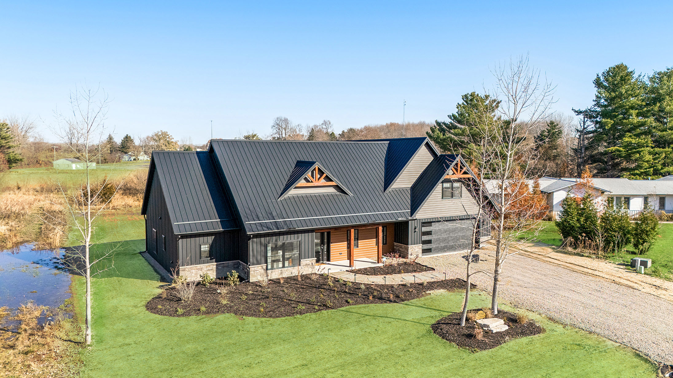 Aerial view of a modern farmhouse-style home with a dark metal roof, dark vertical siding, and natural wood accents, surrounded by a green lawn and some bare trees.