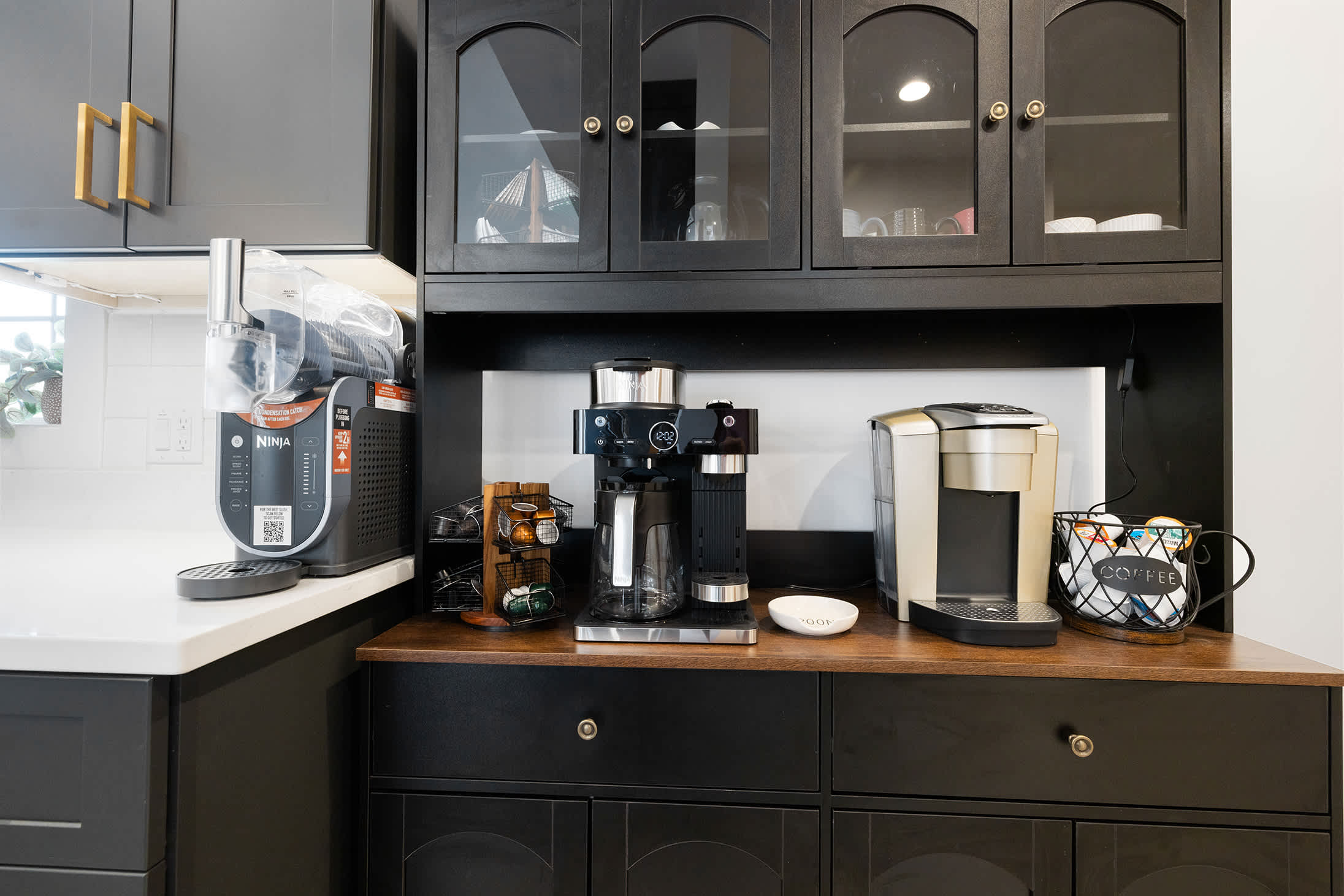 A modern coffee station featuring black cabinetry, a light wood countertop, and various coffee machines including a Ninja and a Keurig, with coffee pod organizers.