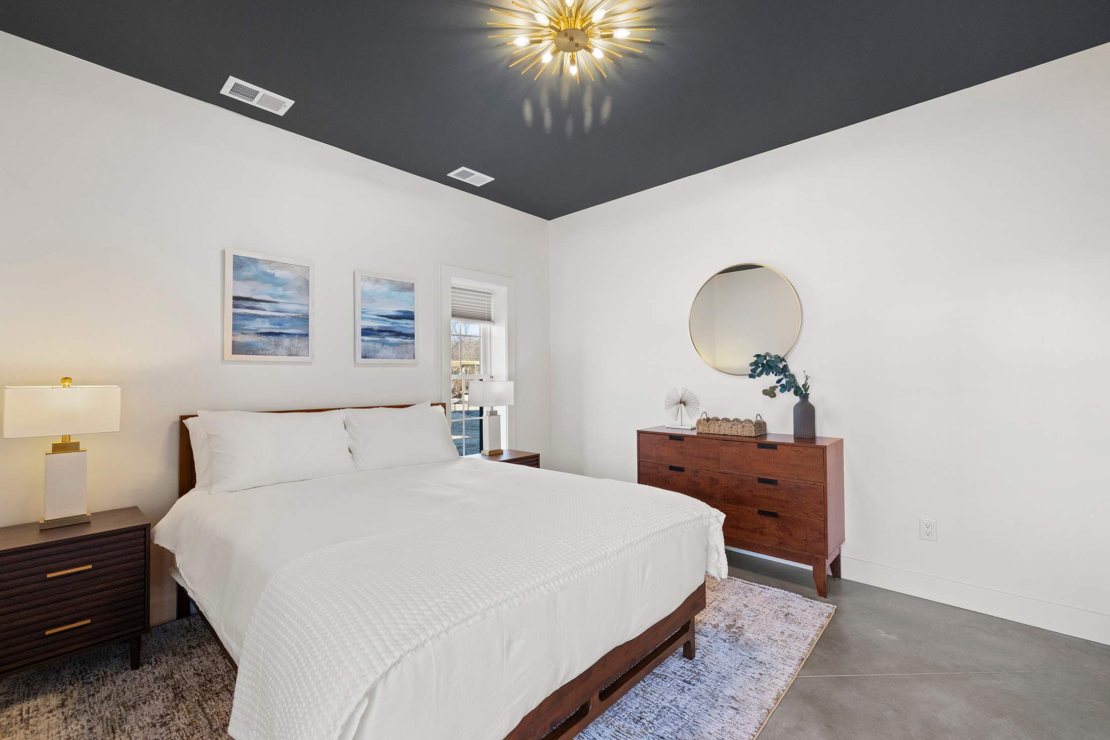 Contemporary bedroom featuring a dark grey ceiling, a queen bed with white linens, a wooden dresser with a round gold-framed mirror, and two windows providing natural light.