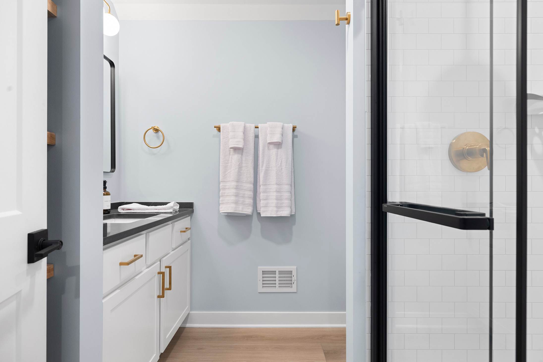 Modern bathroom with light blue walls, a white vanity with a dark countertop, gold hardware, a rectangular mirror, and a glass-enclosed shower with white subway tiles.