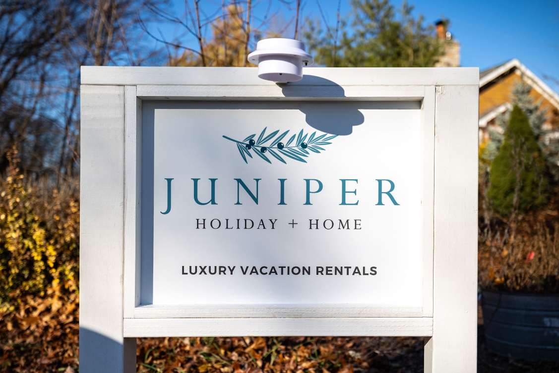 A white wooden sign with 'JUNIPER HOLIDAY + HOME' and 'LUXURY VACATION RENTALS' in blue and black text, set against a backdrop of trees and autumn foliage, with a small white device mounted on top.