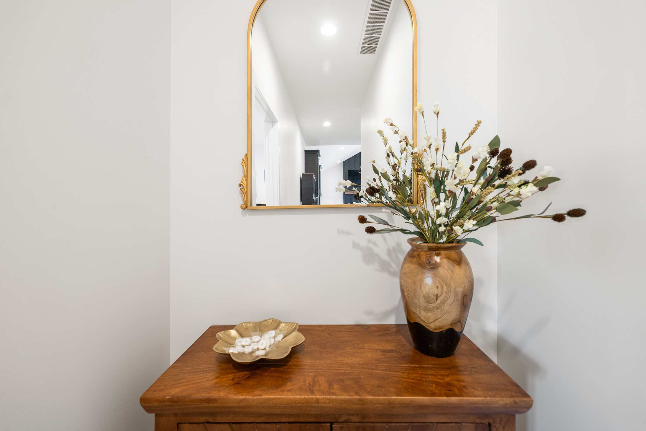 Close-up of an entryway console table with a wooden vase holding dried flowers and a gold decorative bowl, beneath a gold-framed arched mirror reflecting a hallway.
