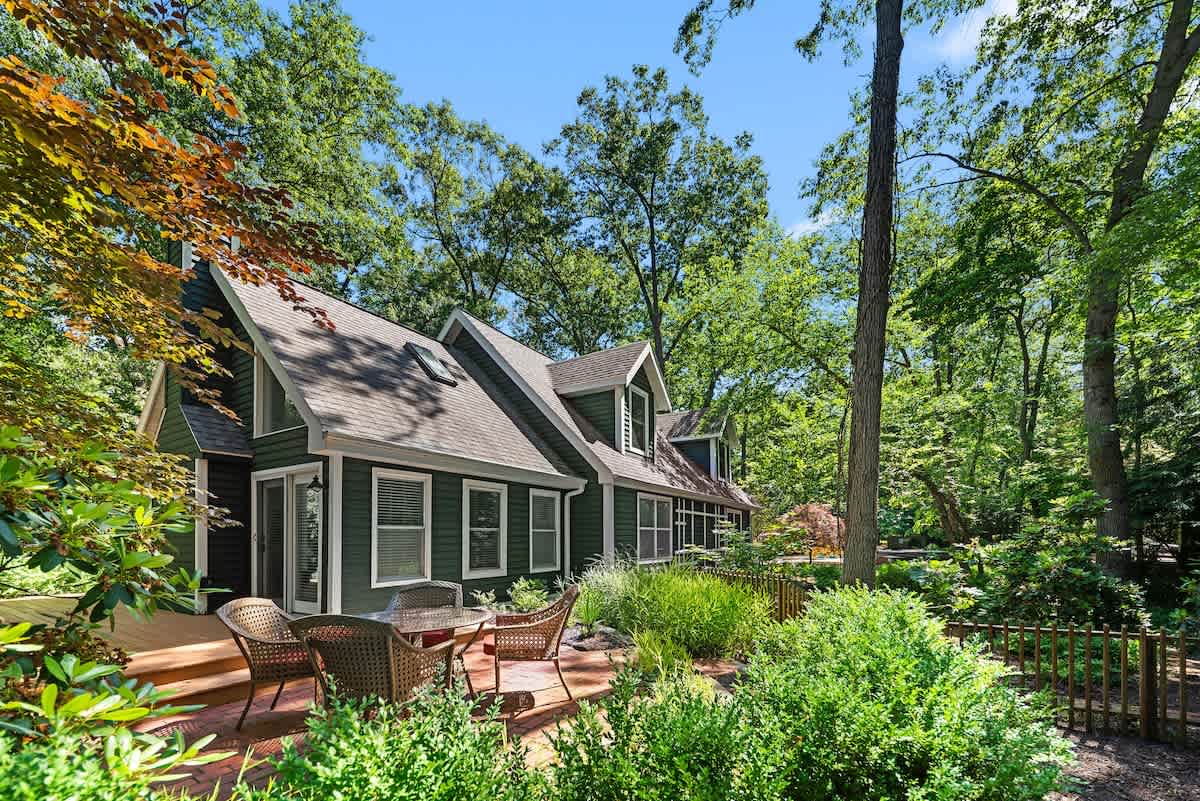 Side view of a dark green house with white trim and multiple windows, nestled among lush trees and greenery. A brick patio with outdoor dining furniture is visible in the foreground, leading to a wooden deck.