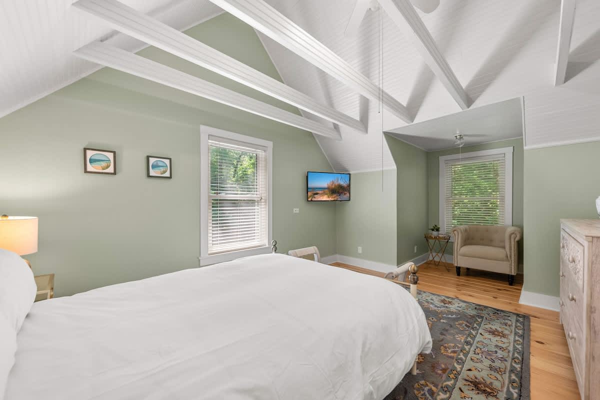 Another view of the cozy bedroom with light green walls, a white bed, and a patterned rug. A wall-mounted TV is visible, along with a window and a small armchair in the corner.