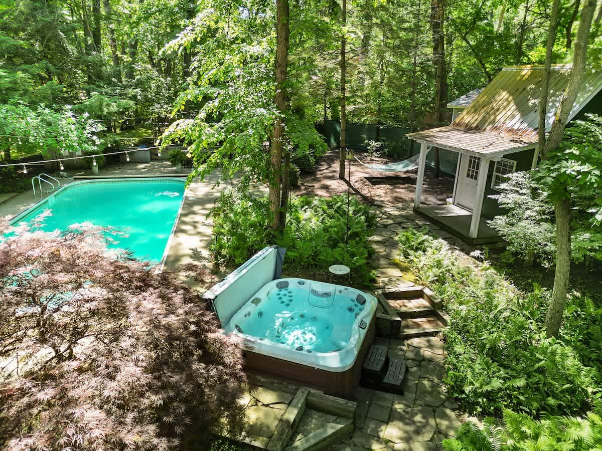 Aerial view of a backyard oasis featuring a sparkling blue swimming pool, a bubbling hot tub with steps, and a small guesthouse or shed, all nestled within a dense forest. Stone pathways connect the amenities, and a hammock is visible in the distance.