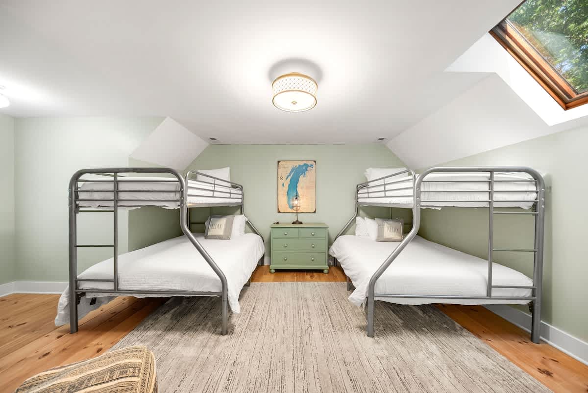 Another view of the bunk room showing two metal bunk beds with white bedding, a green dresser between them, and a skylight. The room has light green walls and a light-colored rug.