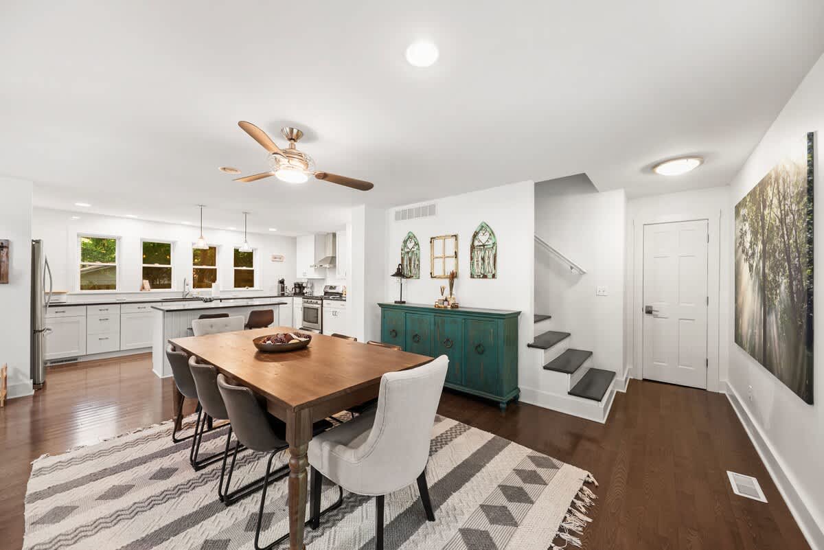 View of the dining room with a wooden table and chairs on a patterned rug, leading towards a staircase with dark treads and a white door. A teal buffet cabinet and large artwork decorate the wall, connecting to the kitchen area.