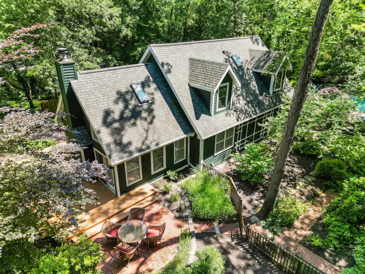 Aerial view of the dark green house with a gray shingled roof and multiple dormer windows, surrounded by a dense canopy of green trees. A wooden deck and brick patio with outdoor furniture are visible below.