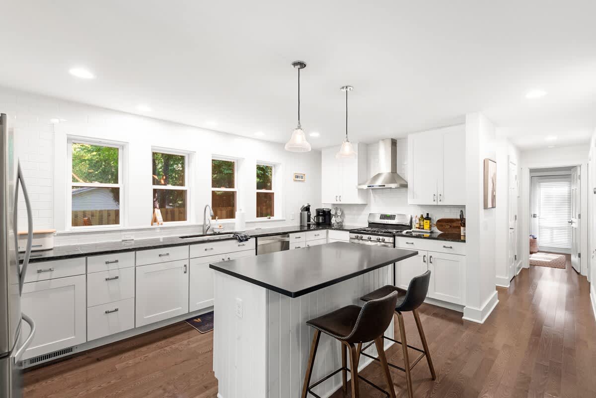 Modern kitchen featuring white shaker cabinets, black countertops, and stainless steel appliances including a gas range and French door refrigerator. A central island with two bar stools and pendant lighting provides additional seating and workspace.