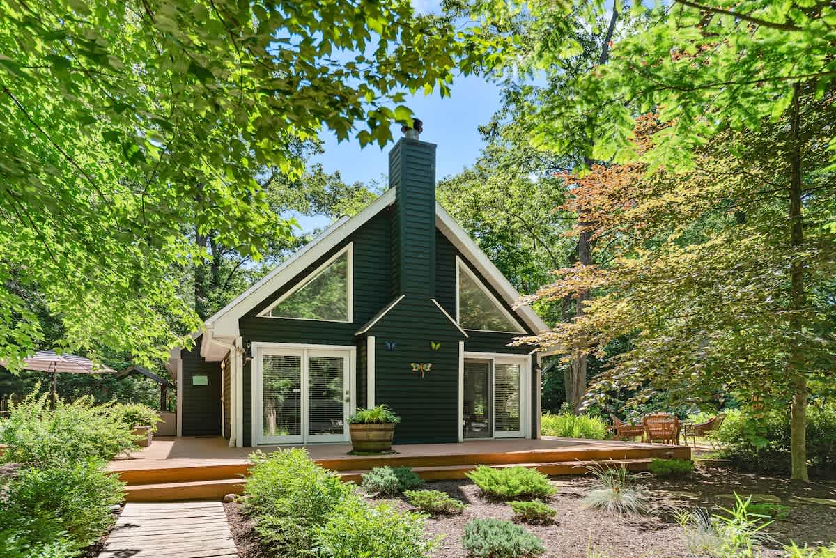 Front view of a charming dark green A-frame style house with a prominent chimney and large triangular windows, surrounded by lush landscaping and mature trees. A wooden deck with outdoor seating extends across the front.